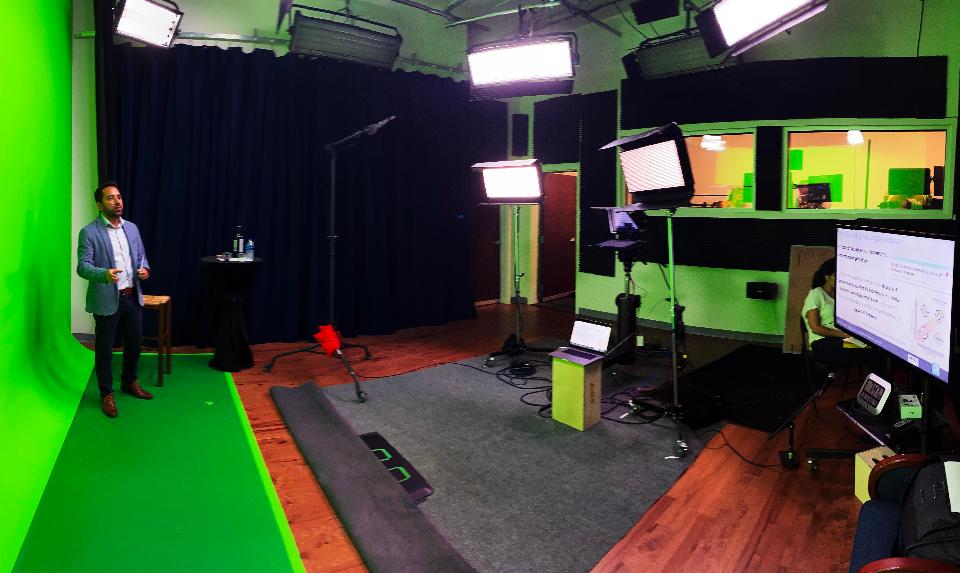 Fig. 6: An advanced studio set up for virtual presentations with additional elements such as a green screen background, multiple cameras and lights, professional microphone, teleprompter, extended monitors and support crew